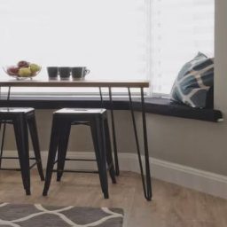 A modern corner breakfast nook with a wooden table and black stools, featuring decorative pillows.