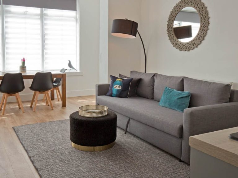Modern living room with a grey sofa, decorative cushions, a floor lamp, and a dining area.