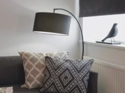 Curved floor lamp beside a sofa with patterned cushions and a small bird figurine on the window.