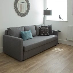 Grey sofa with cushions beside a lamp and window, featuring a decorative mirror on the wall.