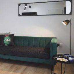 A green velvet sofa with a cushion, a mirror, a floor lamp, and a table with snacks.