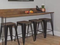 A wooden high table with four black stools, fruit on the table and a plant decoration.